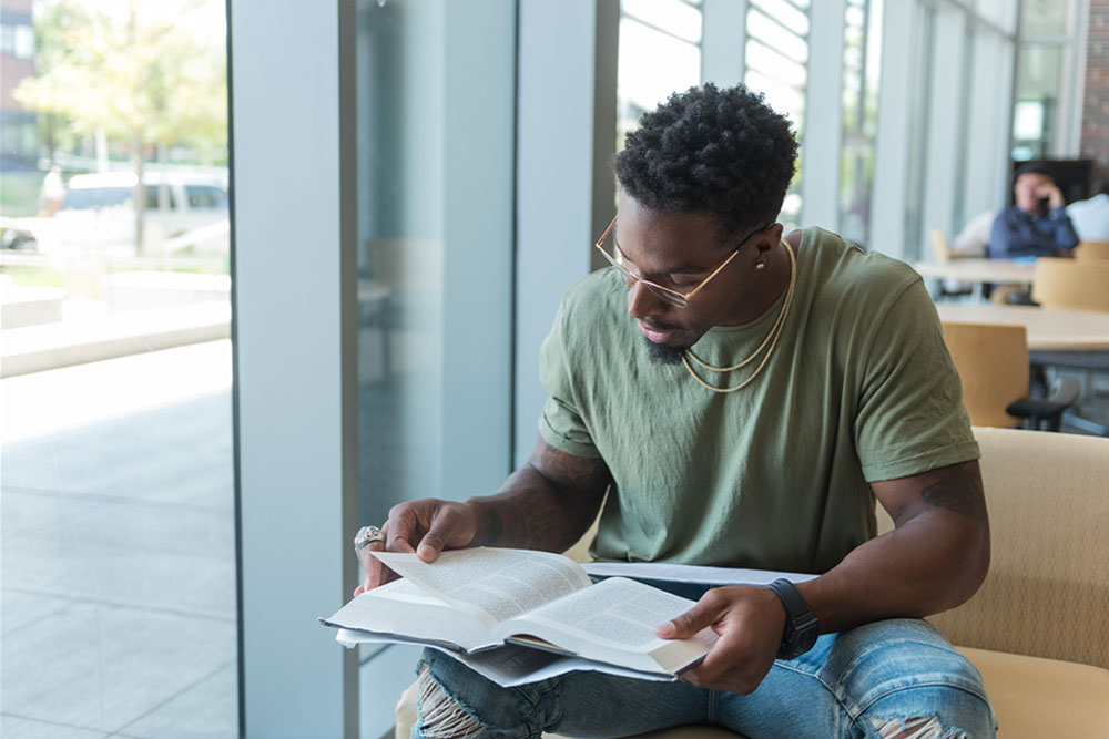 male student reading textbook at bench by window