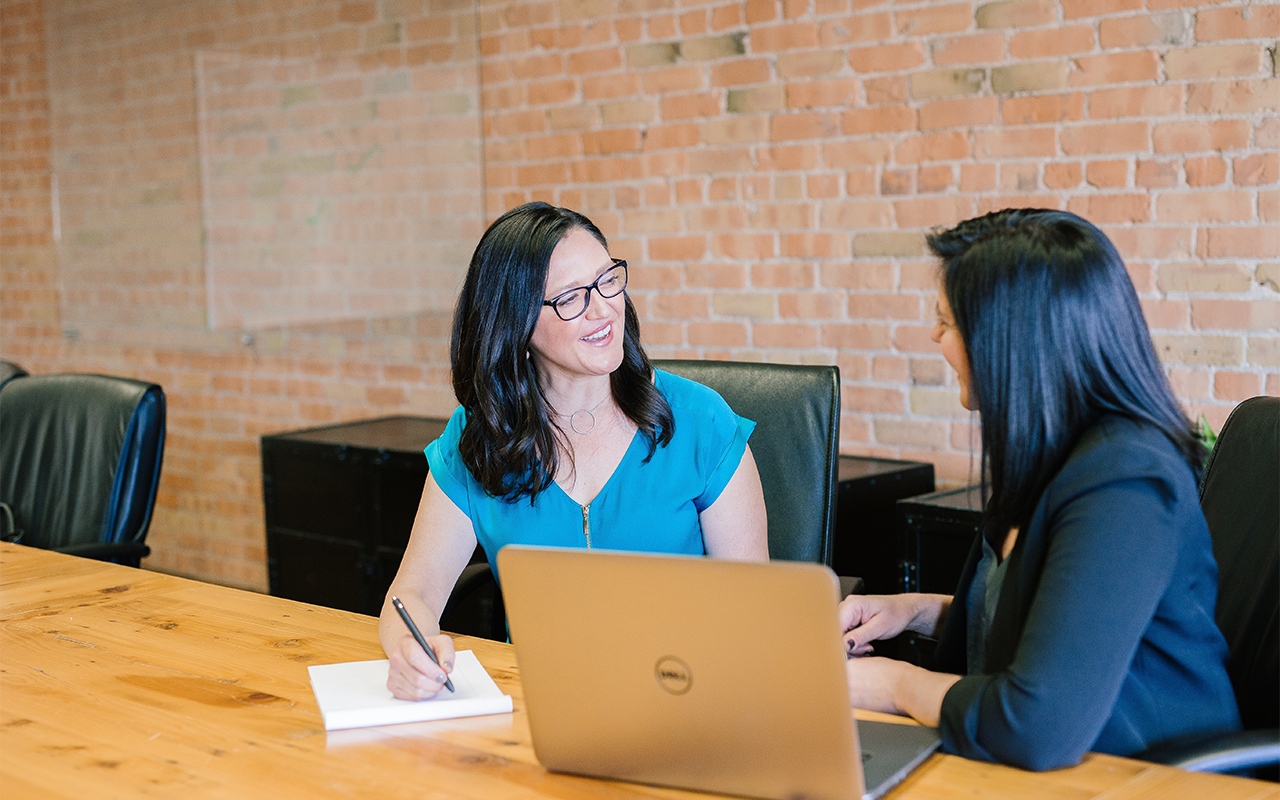 Two women sit side by side at a table, smiling and talking, with a computer and pad of paper in front