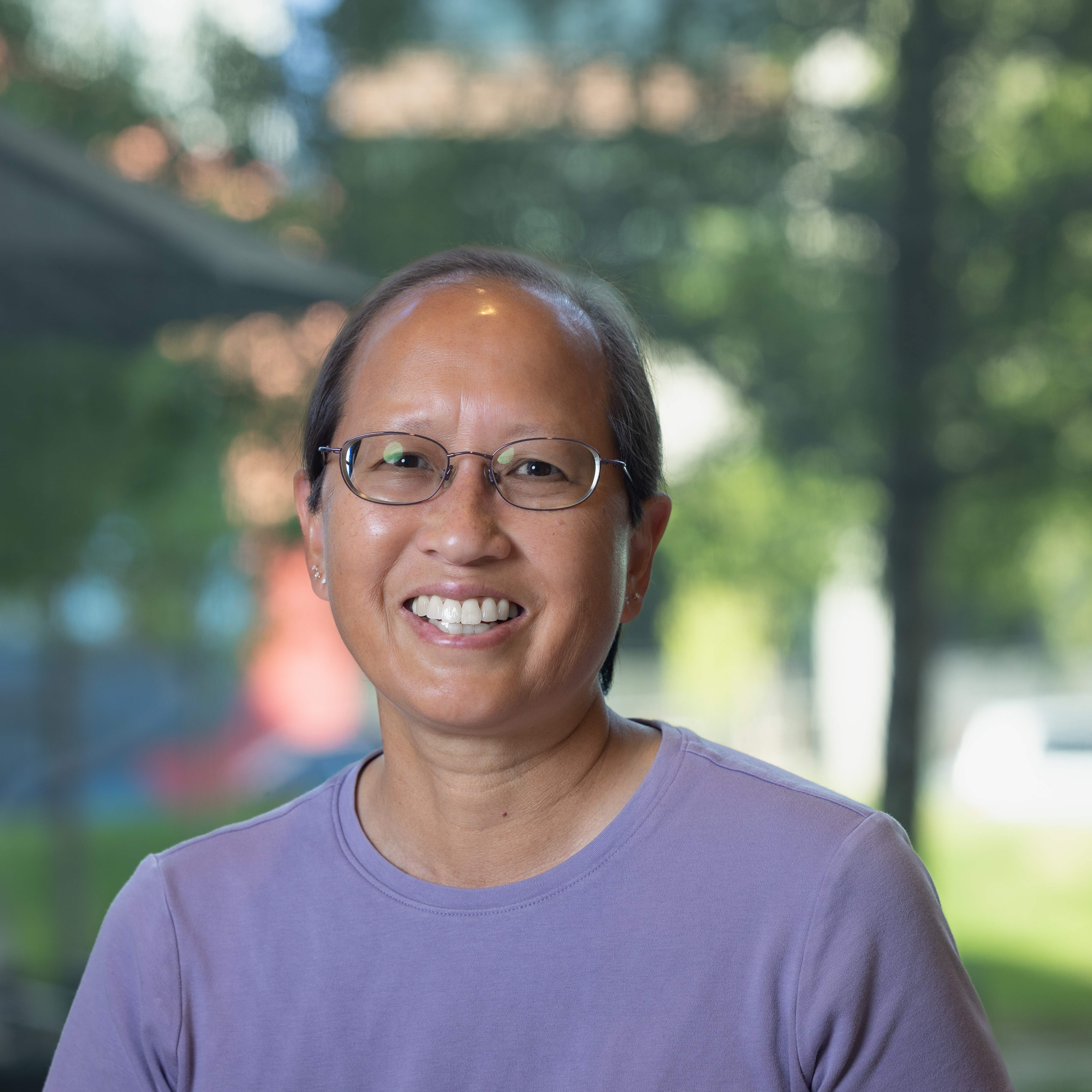 Professional photo of Alice in front of trees. Alice is an Asian American woman with short black hair. Alice is wearing a purple short sleeve shirt and wire rimmed glasses.