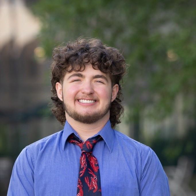 Professional photo of Cayson in front of trees. Cayson is a white man with short, curly, brown hair and a short brown beard. Cayson is wearing a long sleeve blue shirt that is buttoned up and a tie with a red crawfish design.