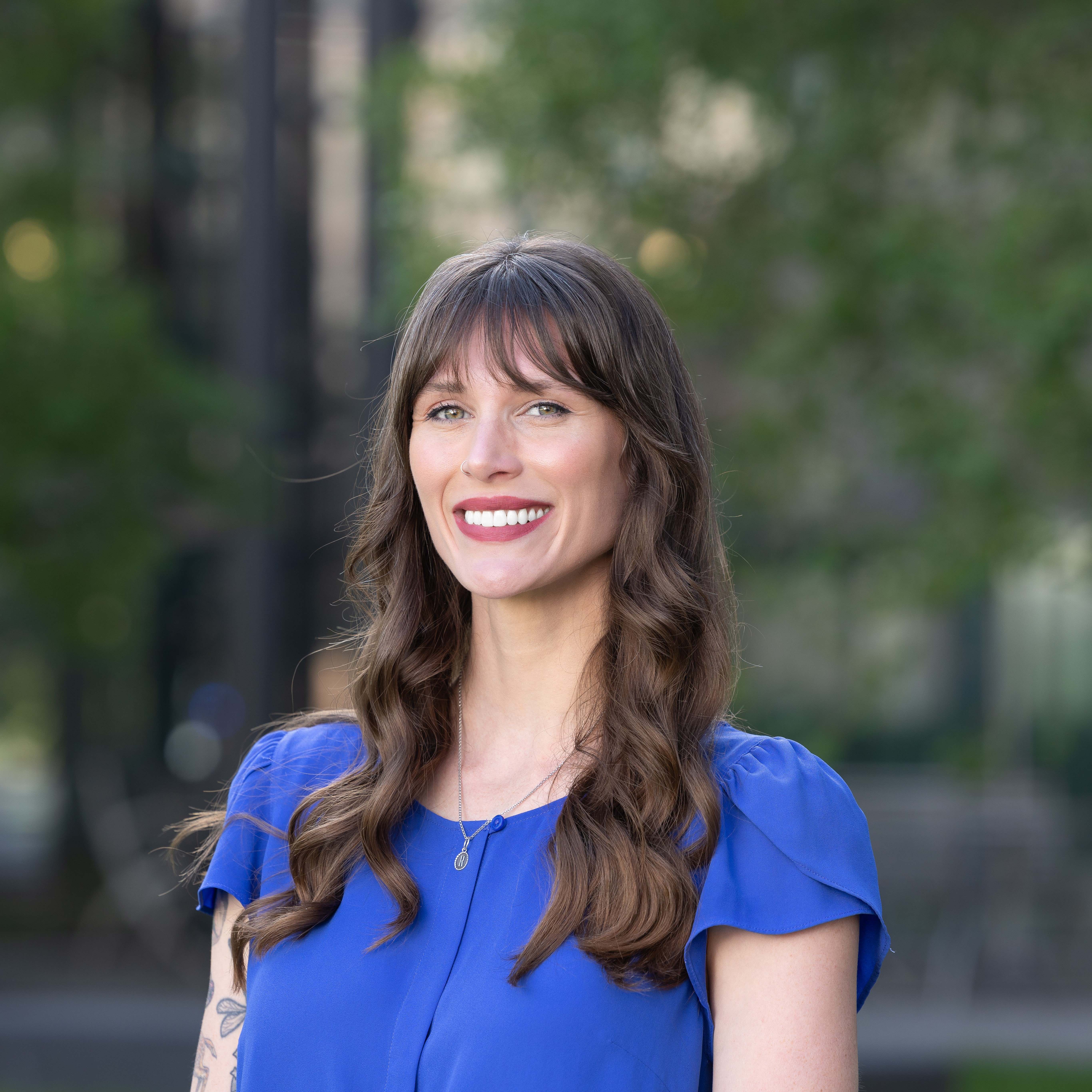 Professional photo of Hanna in front of trees. Hanna is a white woman with long, light brown, curly hair and bangs. Hanna is wearing red lipstick and a royal blue short sleeve shirt.