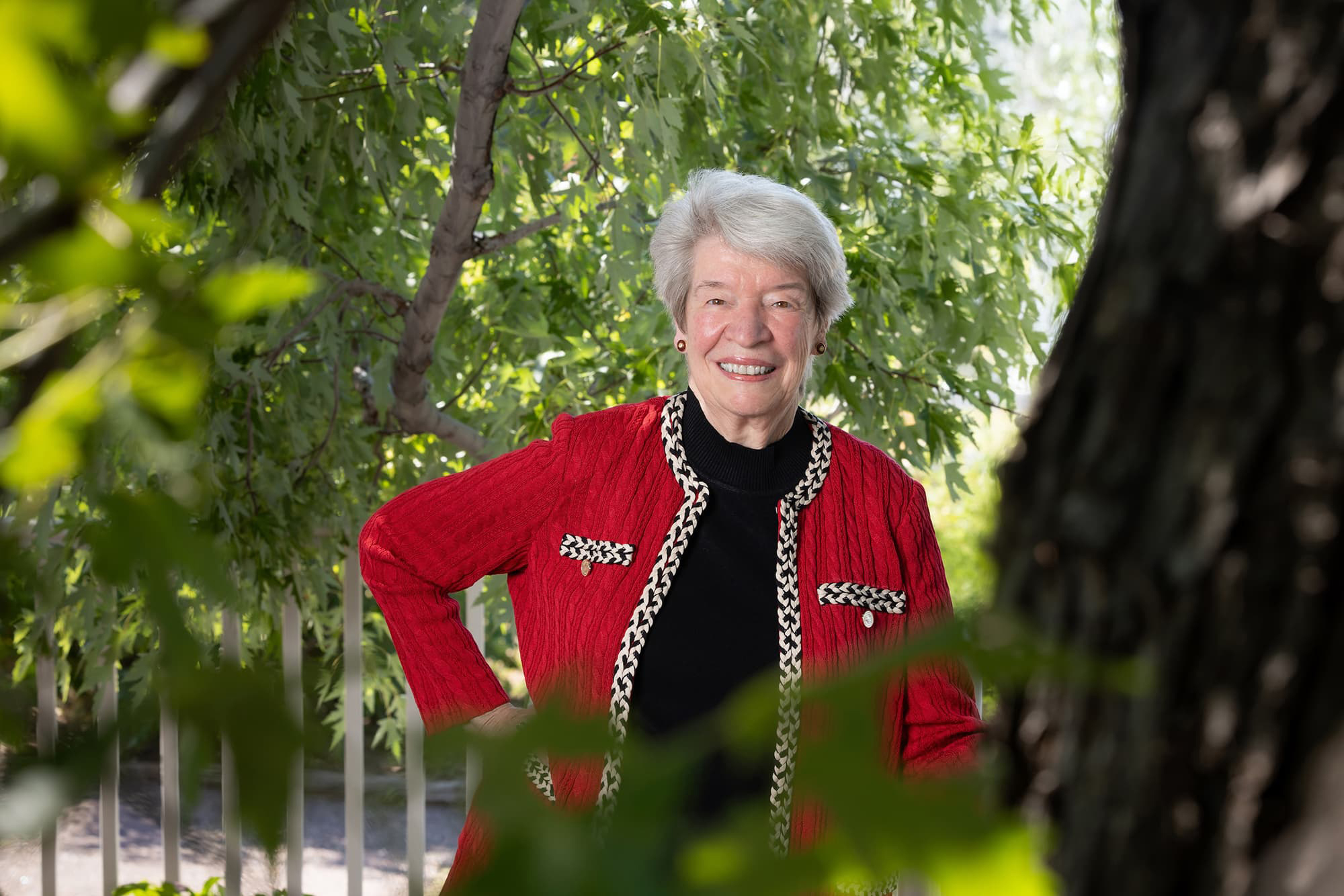 Dr. Mary Guy stands outside amongst trees in a red cardigan and short white hair smiling with her teeth.