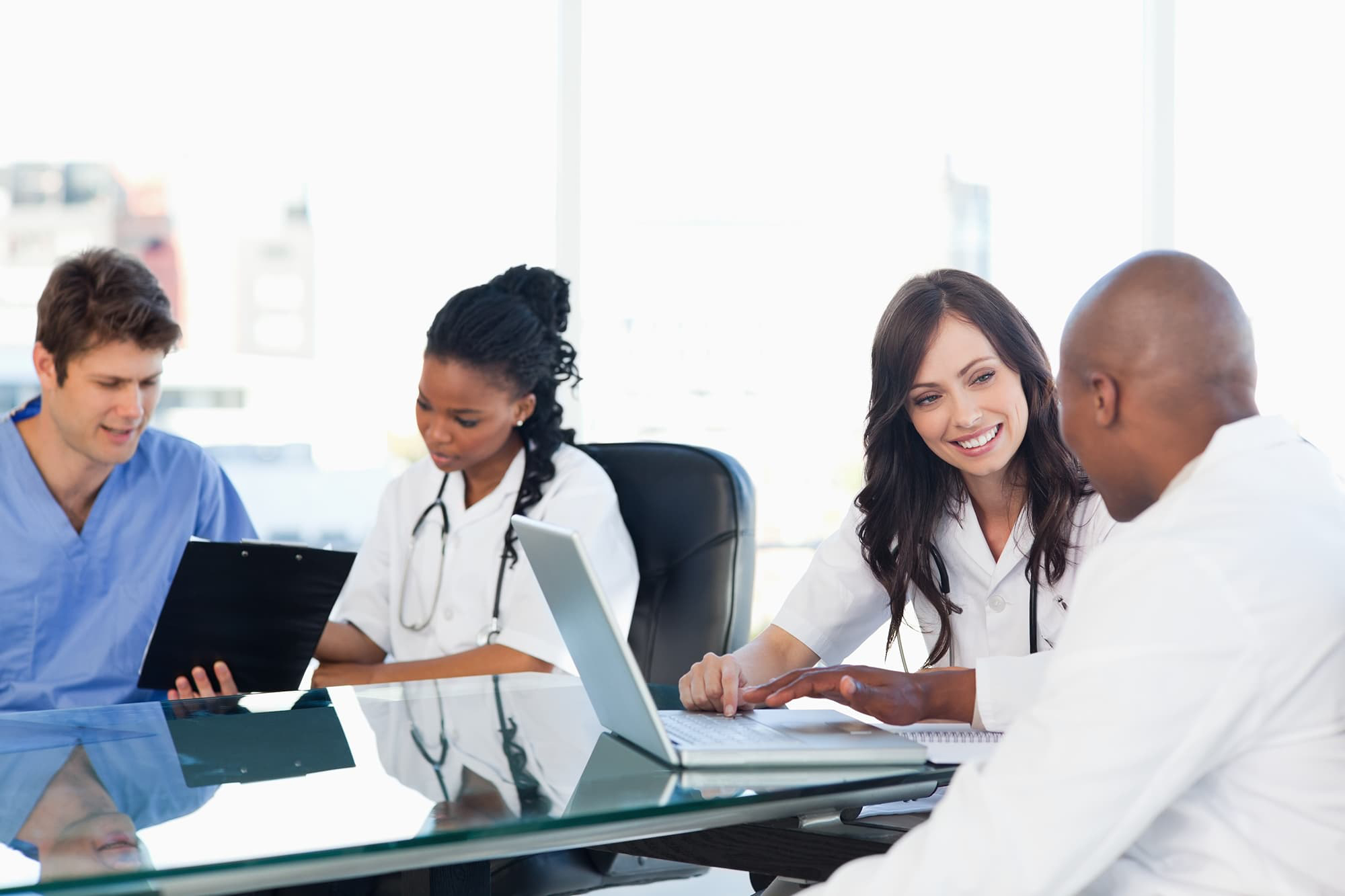 Four medical staff seated at a glass table talking in pairs over laptops.