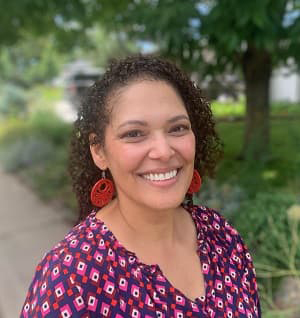 Adrian Lauren Ingram  with dark curly hair, wearing a pink-patterned blouse, smiling with her teeth and standing outside.