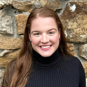 Grace Dugery in a black blouse against a rock-wall backdrop with long hair, smiling with her teeth.