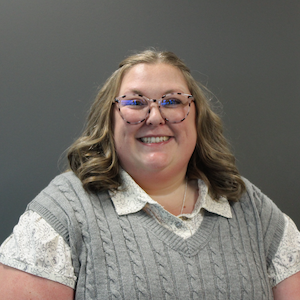 Woman with shoulder-length dark blond hair and turtle shell glasses in a gray sweater smiling with her teeth.