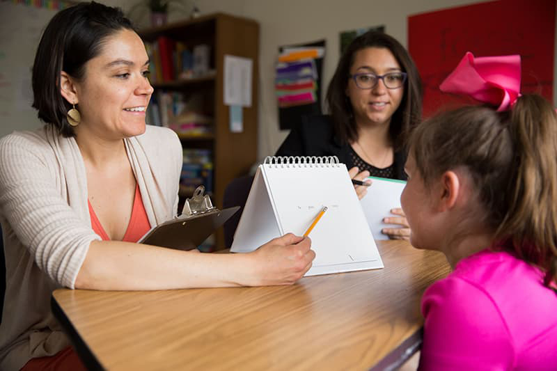 Two teachers working with a young student at a desk, all smiling.