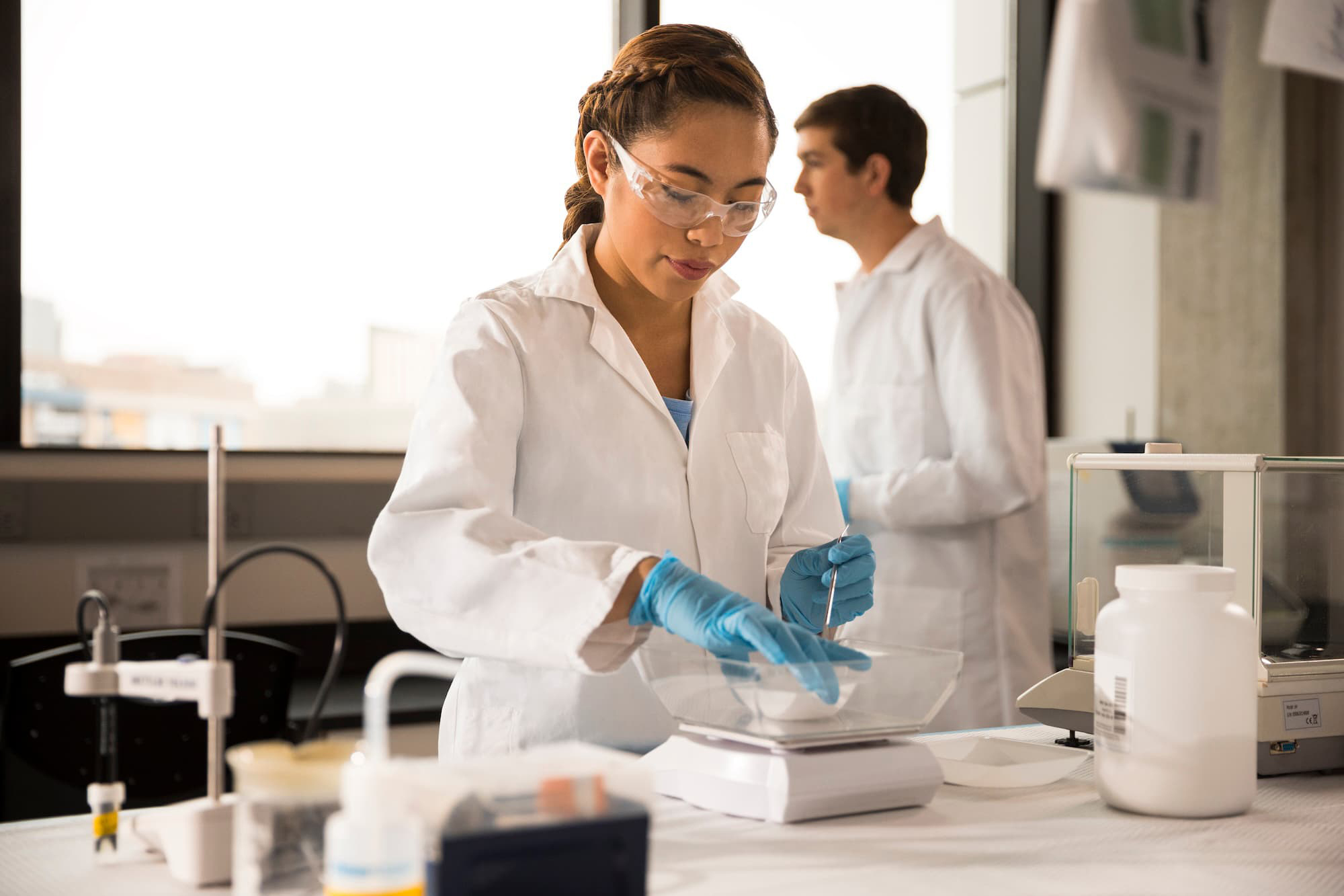 Student wearing a white lab coat, blue gloves, and safety glasses working in a lab.