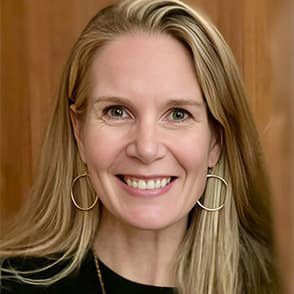 Alexa Braun with long blonde hair in a dark blouse and gold hooped earrings, smiling with her teeth.