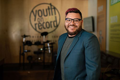 Andy Guerrero with pink hair and a blue blazer smiles with his teeth, wearing square dark-rimmed glasses.