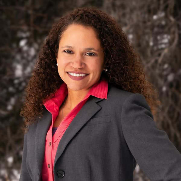 Headshot of Brandy Reitter in a gray blazer and bright pink button down shirt, dark curly, long hair, and smiling with her teeth.