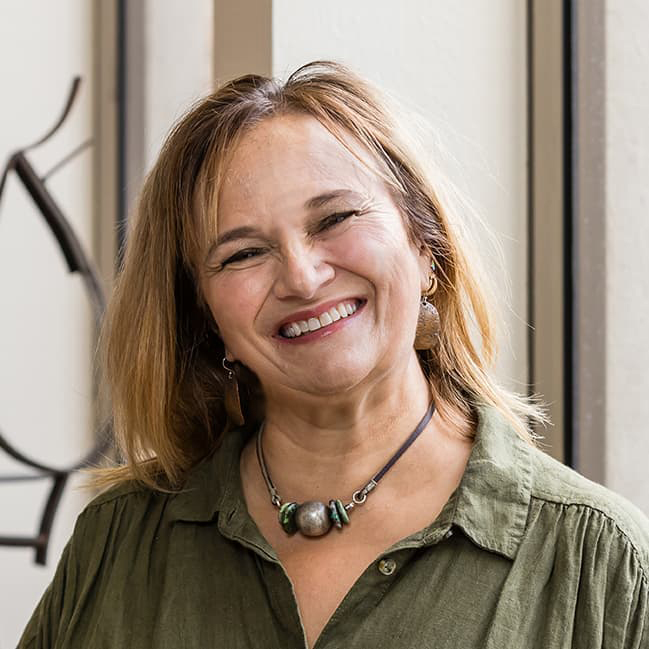 Helen Douglass in a forest green blouse and chunky jewelry with shoulder length blonde hair smiles with her teeth.